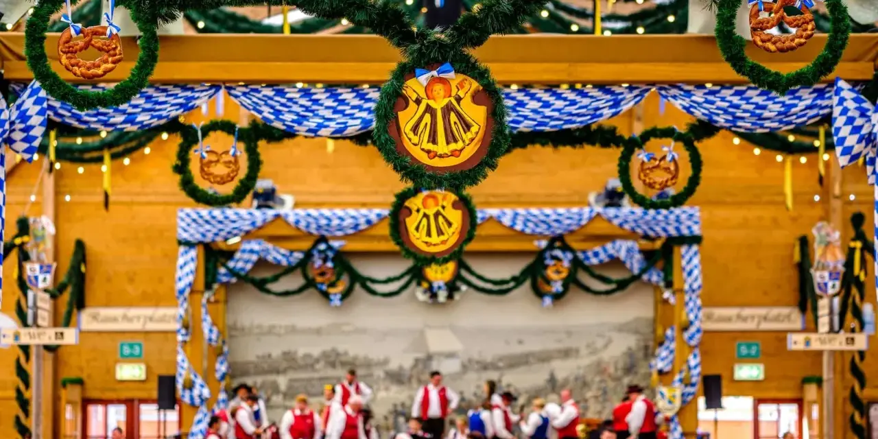 Wnętrze namiotu na Oktoberfest ozdobione niebiesko-białymi girlandami i wiankami z precelkami. Ludzie siedzą przy stołach, a w tle widać orkiestrę.