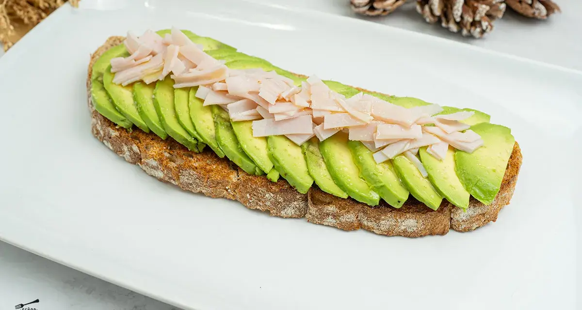 Tostada integral con aguacate en rodajas y jamón picado sobre plato blanco.