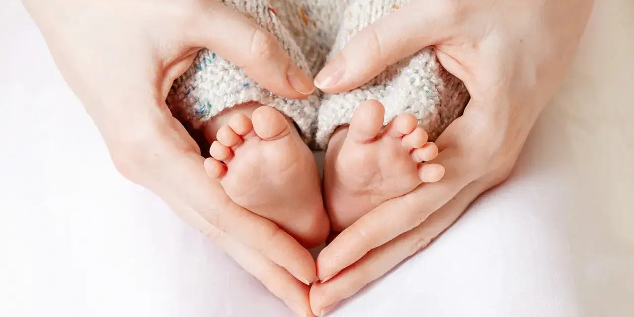 Manos formando un corazón alrededor de los pies de un bebé, evocando la protección y el complemento de maternidad con retroactividad.