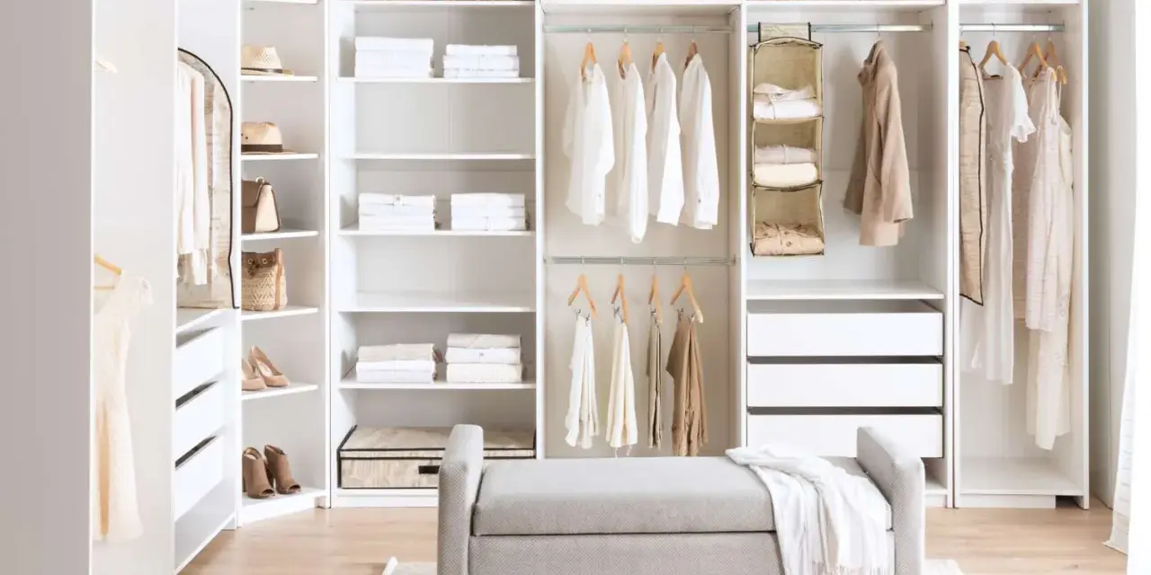 A bright, organized closet with white DIY closet shelves, featuring neatly folded clothes, baskets, and hanging garments. A grey bench sits in front.