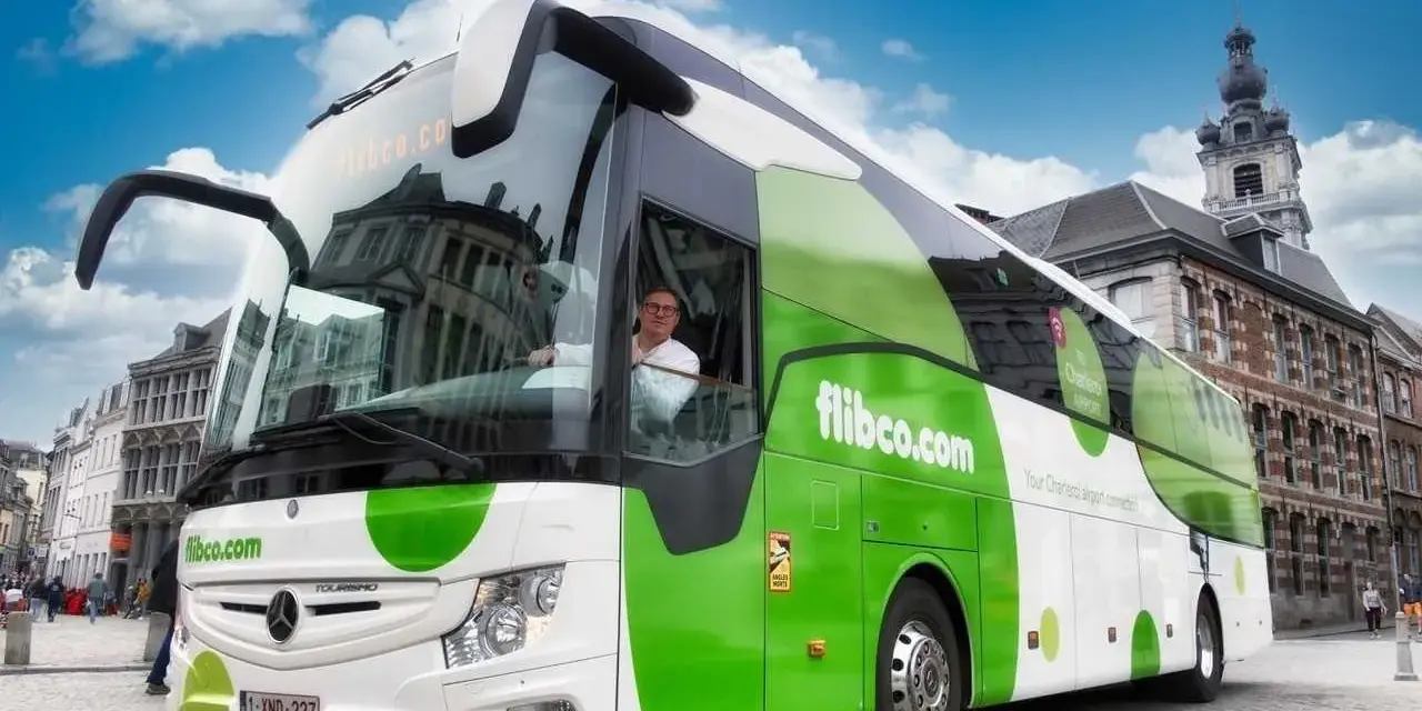 Autobus flibco.com z kierowcą, na tle miejskiej zabudowy i błękitnego nieba.