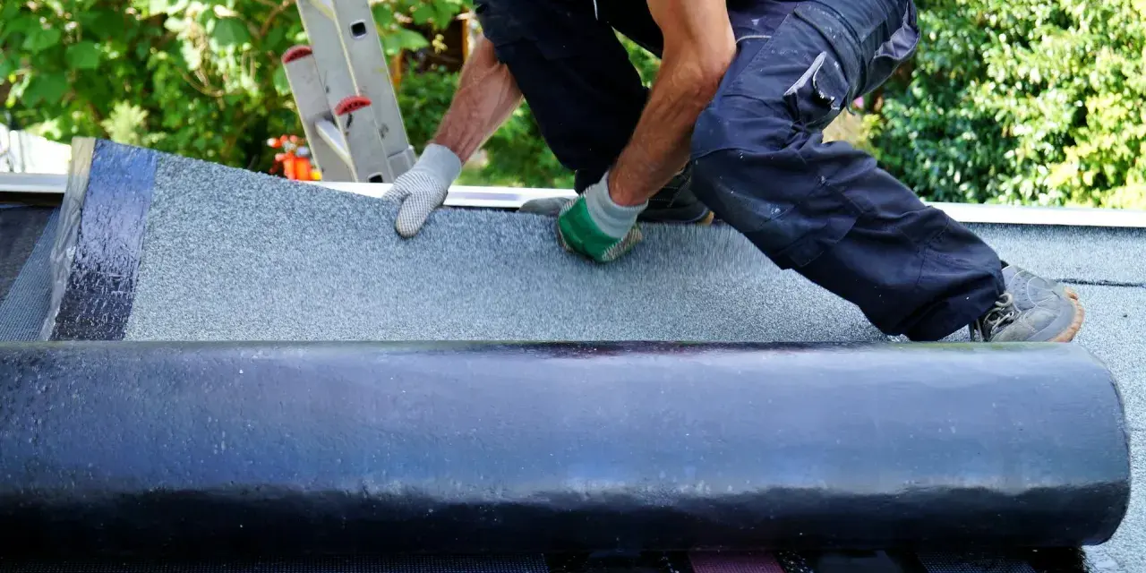 Handwerker verlegt Dachschindeln. Eine Rolle Bitumenbahn liegt bereit, während die neue Schicht auf dem Dach ausgebreitet wird.