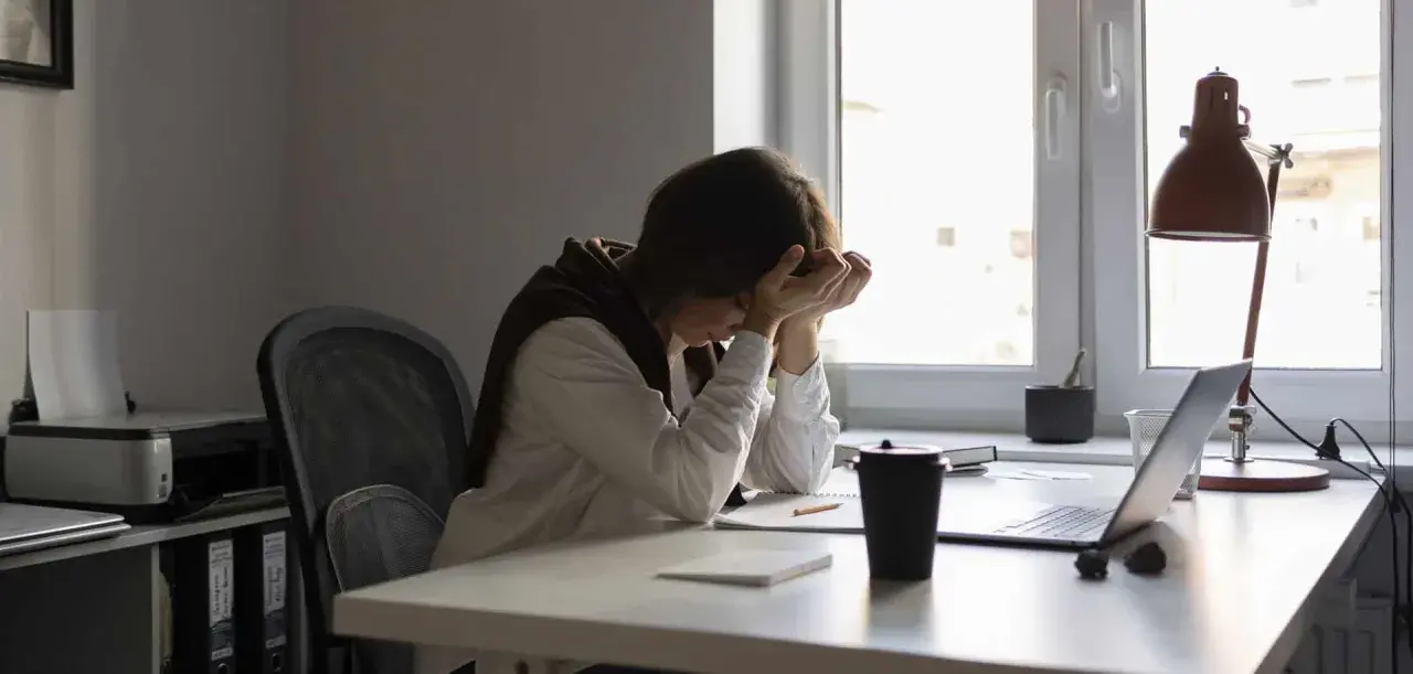 Une personne accablée par la pression au travail, la tête entre les mains, assise à un bureau encombré.