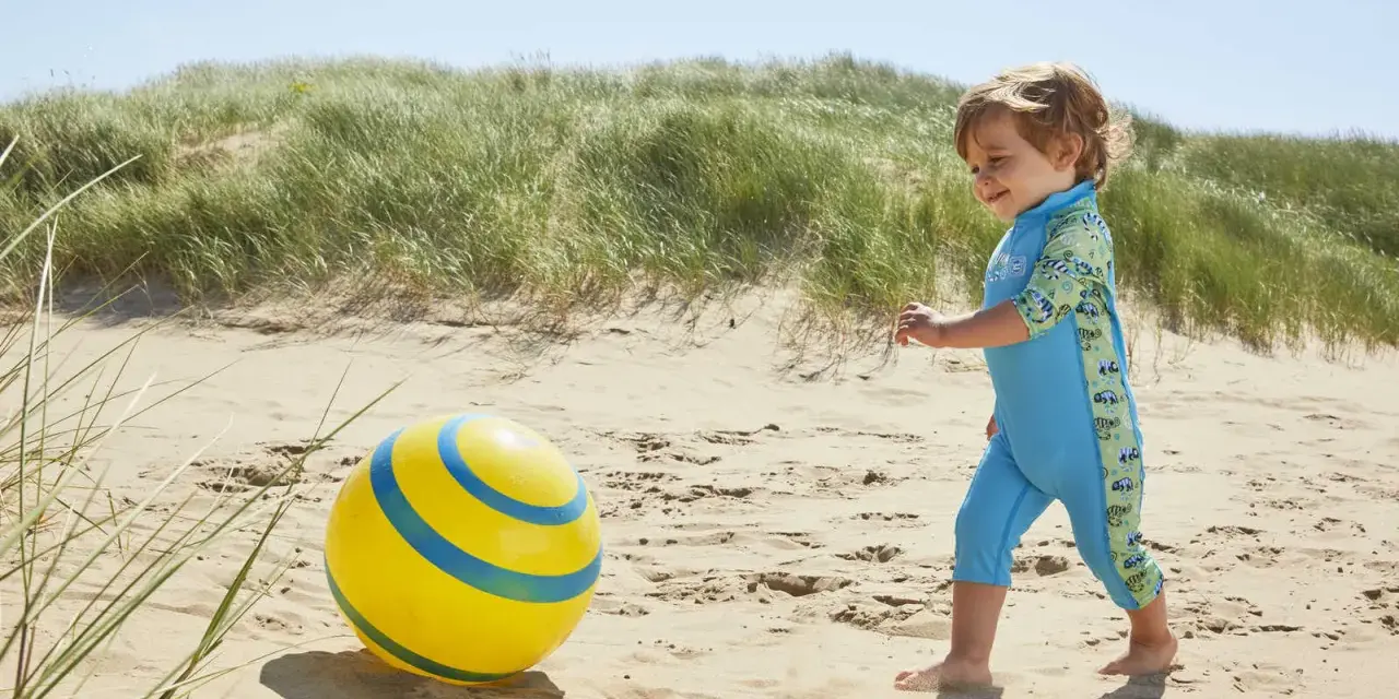 Chłopiec w niebieskim stroju kąpielowym z neoprenu bawi się żółtą piłką na plaży.