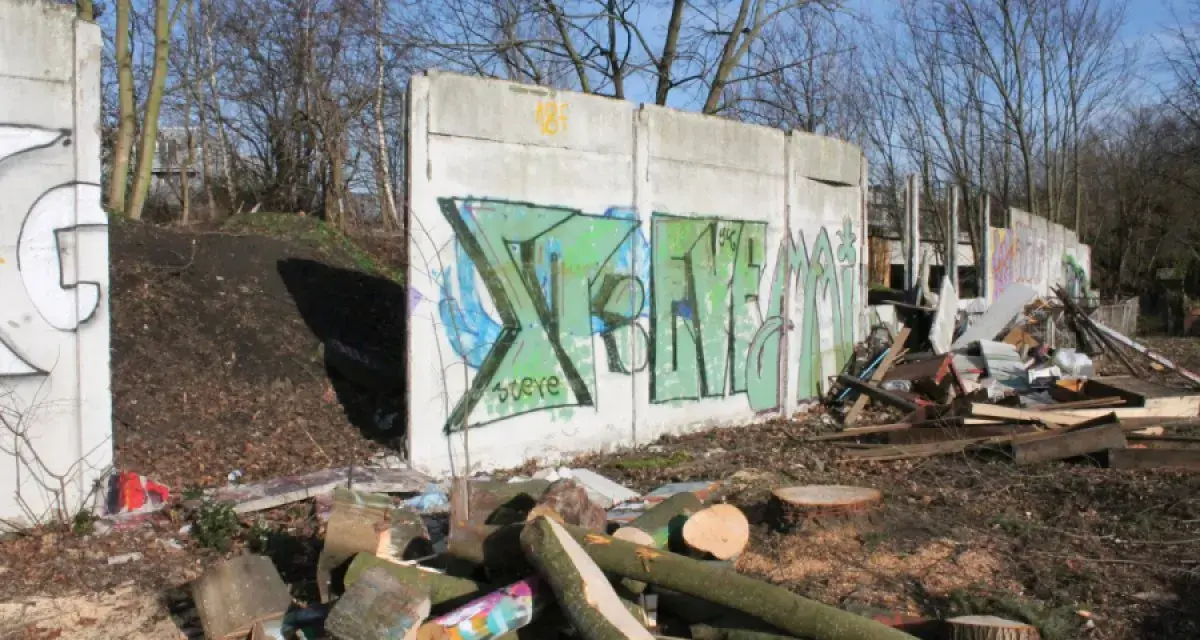 Graffiti-bemalte Überreste der Berliner Mauer, umgeben von Holzstapeln und Trümmern.