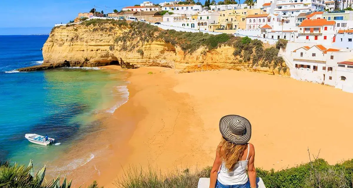 A woman in a sun hat enjoys the view of a golden beach and turquoise water in Portugal's Algarve region.