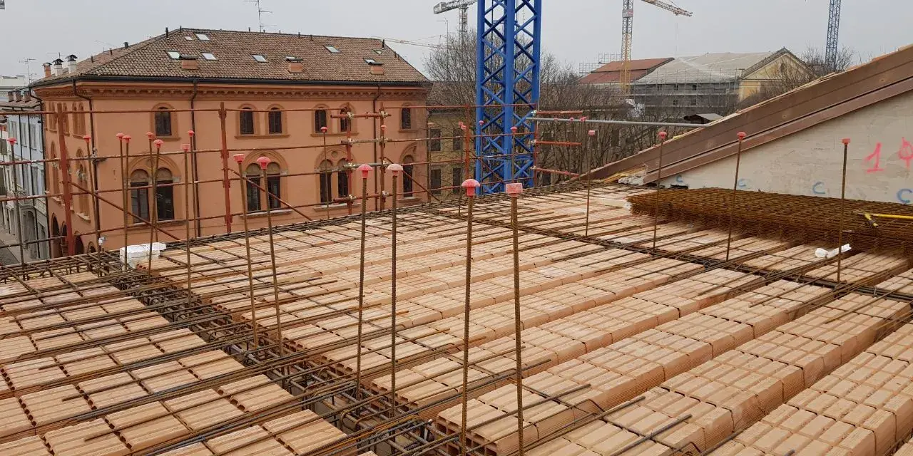Costruzione di un solaio: mattoni forati e armature in ferro pronti per il getto di cemento. In lontananza, un edificio storico e gru edili.
