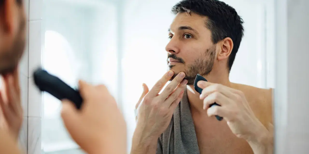 Hombre con barba corta se afeita la barba con una recortadora, buscando un afeitado apurado frente al espejo.