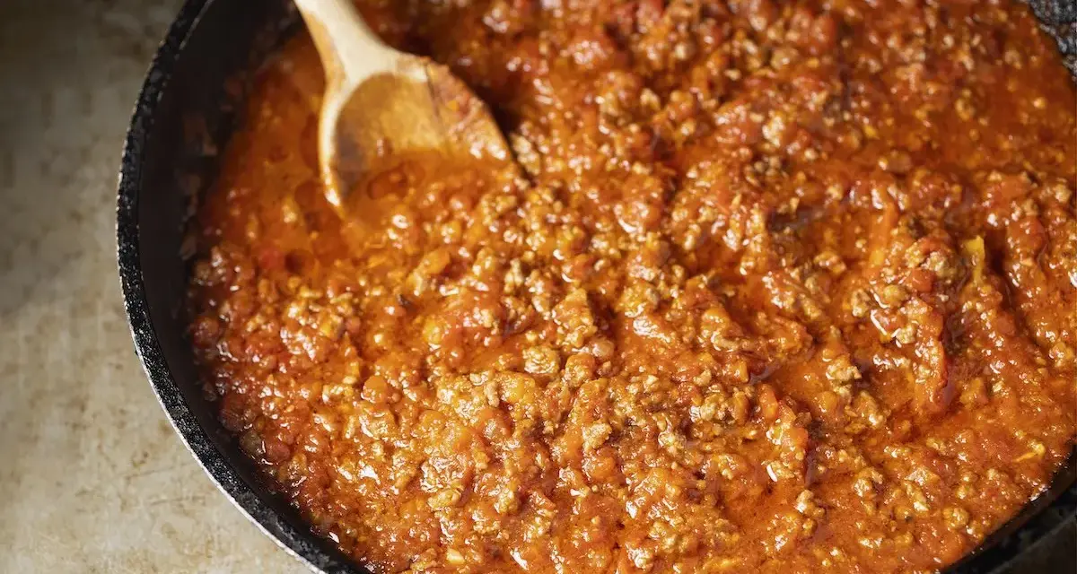 Un saporito ragù di carne in una padella, pronto per essere servito. Un cucchiaio di legno mescola la salsa ricca e densa.