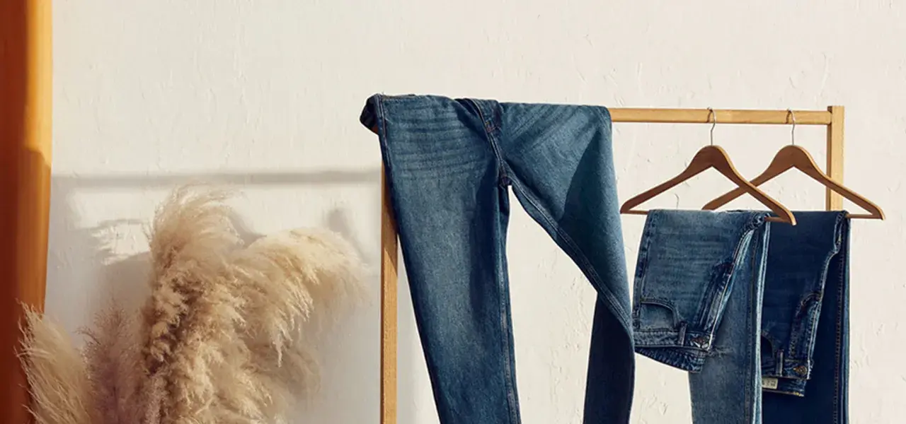 Varios pares de pantalones de **tejido denim** cuelgan de una percha de madera. A la izquierda, un gran ramo de hierba de la pampa añade un toque natural.