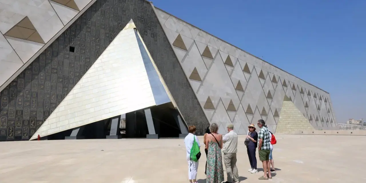 Grupa turystów podziwia nowoczesne muzeum w Gizie, którego fasada zdobiona jest hieroglifami i geometrycznymi wzorami przypominającymi piramidy.