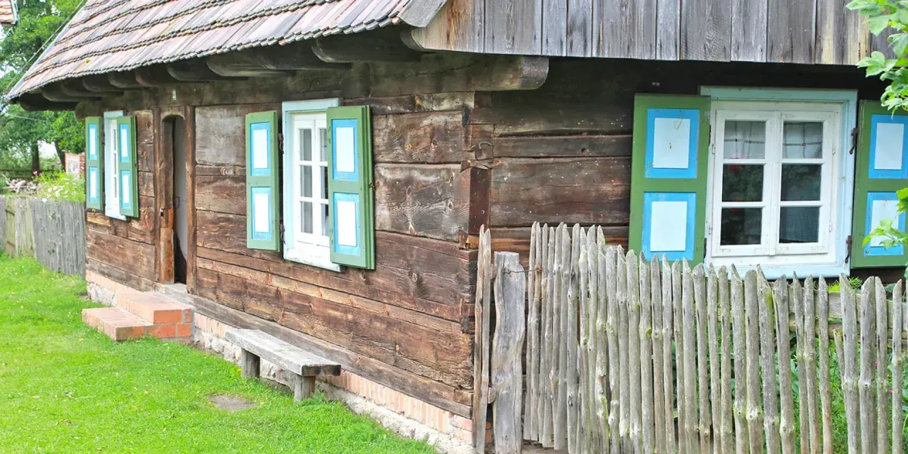 Drewniana chata z zielono-niebieskimi okiennicami, ławką i płotem. Skansen w Dziekanowicach zaprasza do podróży w czasie.