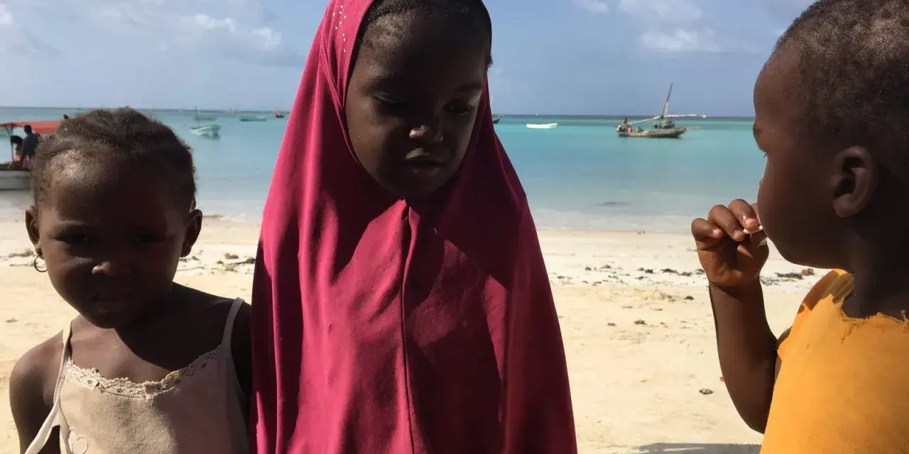 Dzieci na plaży, w tle turkusowe morze i łodzie. To obrazek z życia na Zanzibarze.