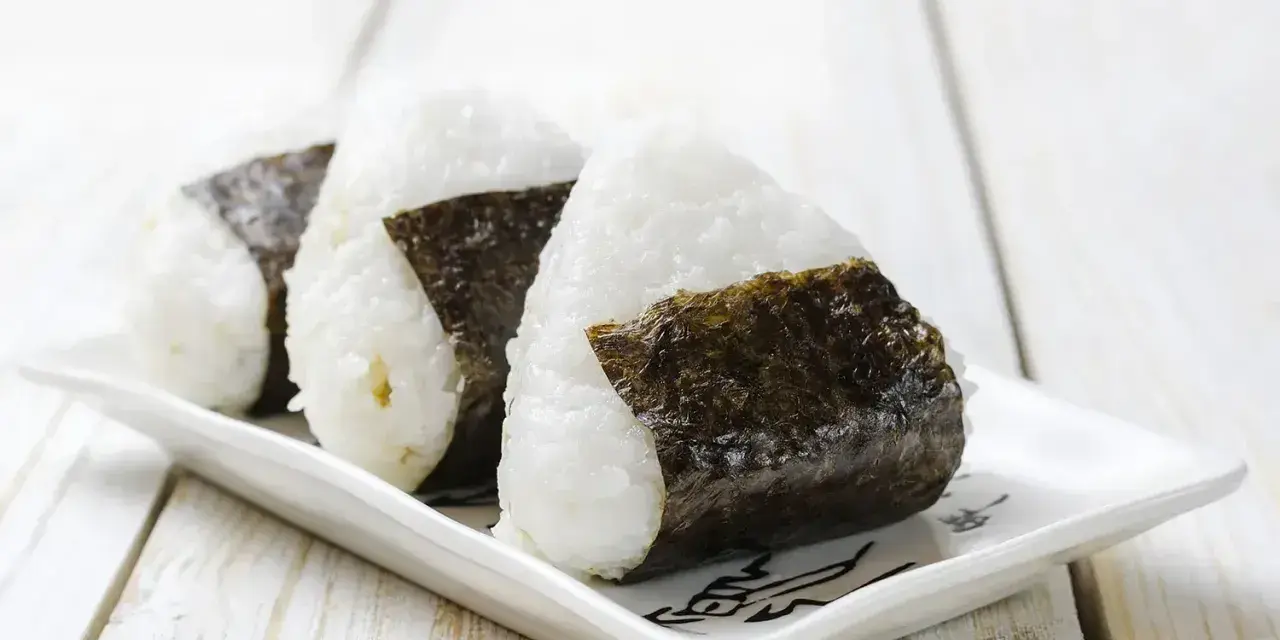 Trzy japońskie kulki ryżowe, onigiri, owinięte w ciemne wodorosty nori, ułożone na białej, ozdobnej tacce.