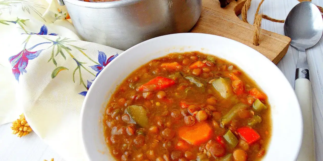 Guiso de lentejas con verduras en un cuenco blanco, junto a una olla y servilleta floral.