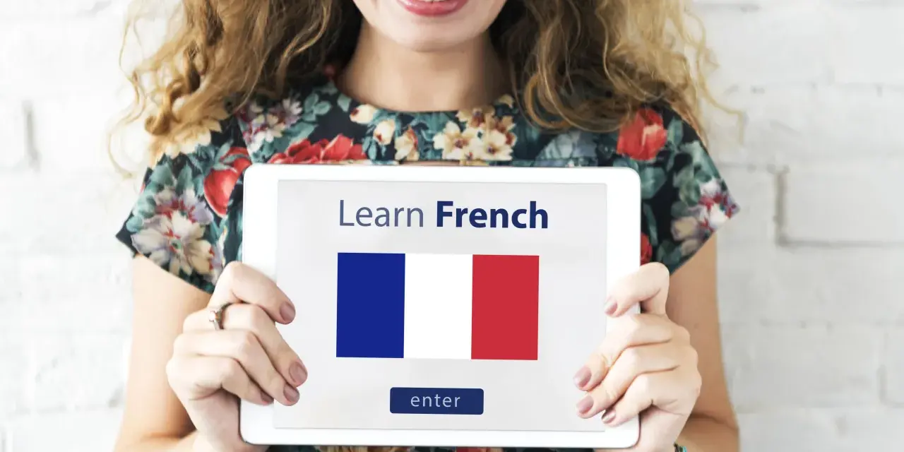 Una mujer sonriente sostiene una tablet que muestra "Learn French" y la bandera francesa. ¡Es hora de aprender como se habla en Francia!