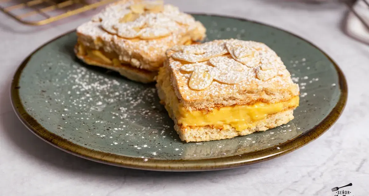 Dos porciones de pastel ruso con crema amarilla y almendras laminadas, espolvoreados con azúcar glas.