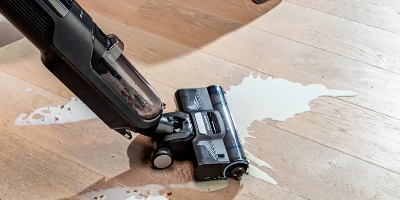 A wet/dry vacuum cleaner is cleaning up a spill of white liquid and debris on a wooden floor.
