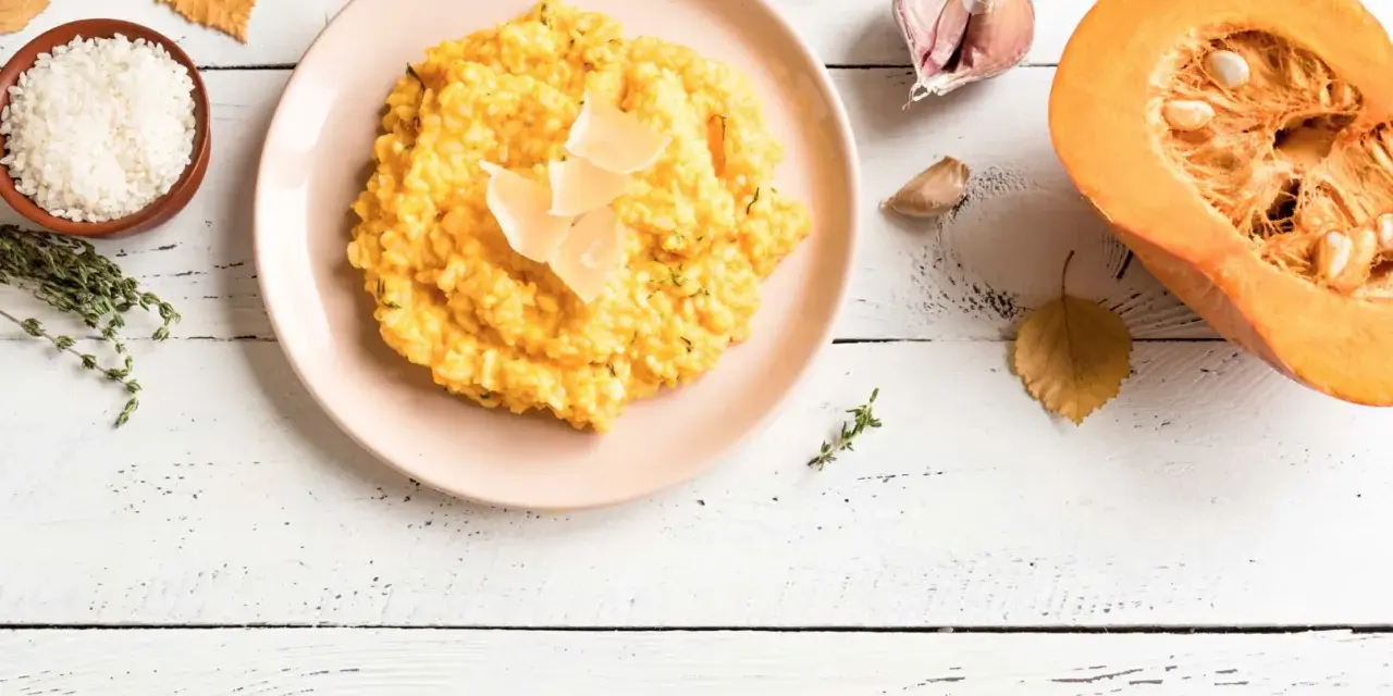 Un cremoso risotto de calabaza, decorado con láminas de queso, junto a ingredientes frescos como arroz, tomillo, ajo y una calabaza partida.