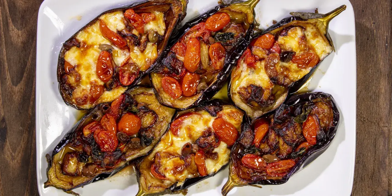 Melanzane a scarpone ripiene di pomodorini, formaggio filante e olive, cotte al forno su un piatto bianco.