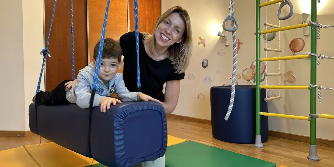 Chłopiec z ADHD u niemowlaka ćwiczy na huśtawce terapeutycznej z uśmiechniętą fizjoterapeutką.