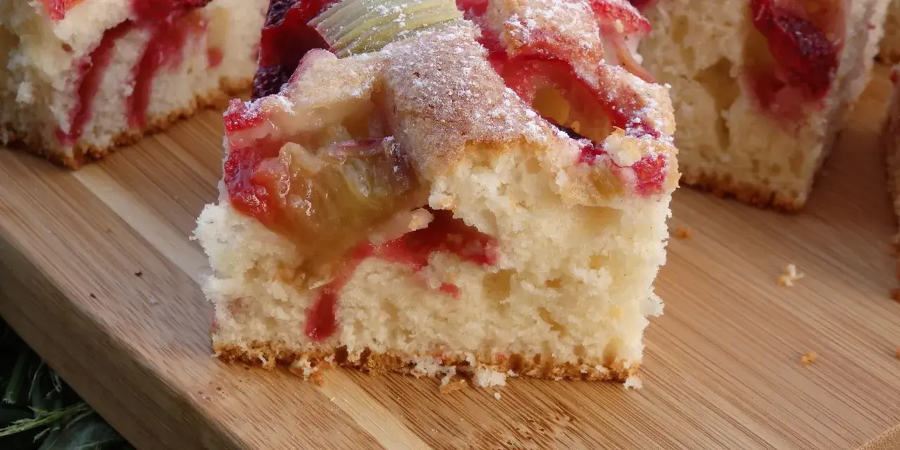 Puszyste ciasto na kefirze z rabarbarem i truskawkami, posypane cukrem pudrem, na drewnianej desce.