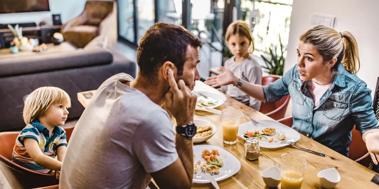 Familie am Esstisch, die Frau erklärt gestikulierend, wie streitet man richtig, während der Mann sich die Ohren zuhält.