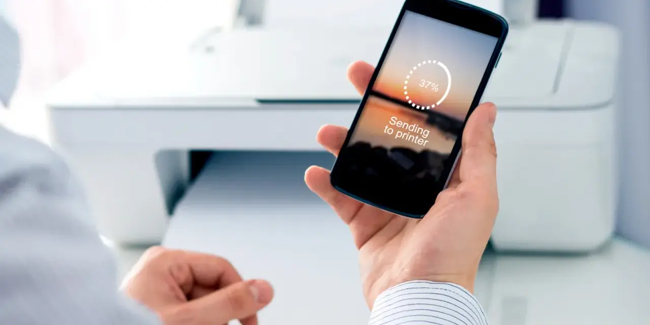 Dłoń trzyma smartfon z napisem "Sending to printer", pokazując jak drukować z telefonu. W tle biała drukarka.