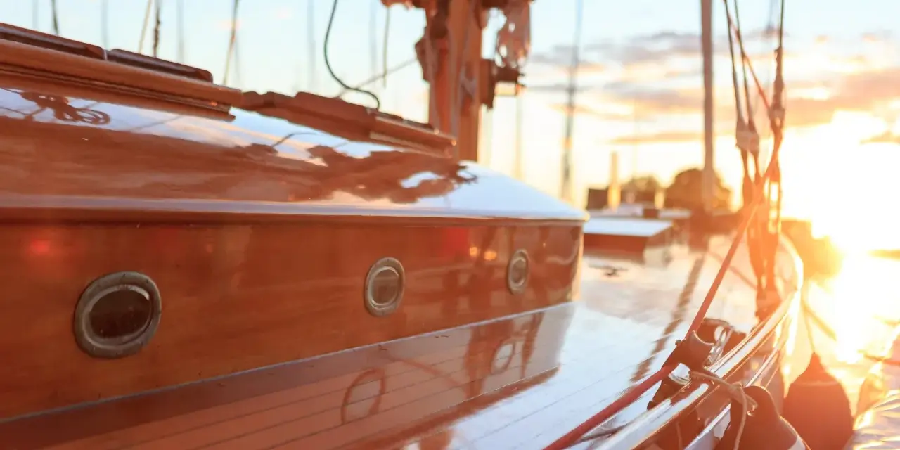 Glänzendes Holzdeck eines Segelboots, frisch lackiert. Kleine Bullaugen spiegeln die untergehende Sonne wider.