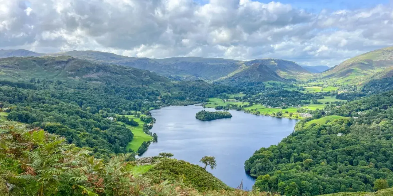 A stunning view of a lake surrounded by green hills and trees, perfect for day trips from Liverpool. A lone hiker walks down a path in the foreground.