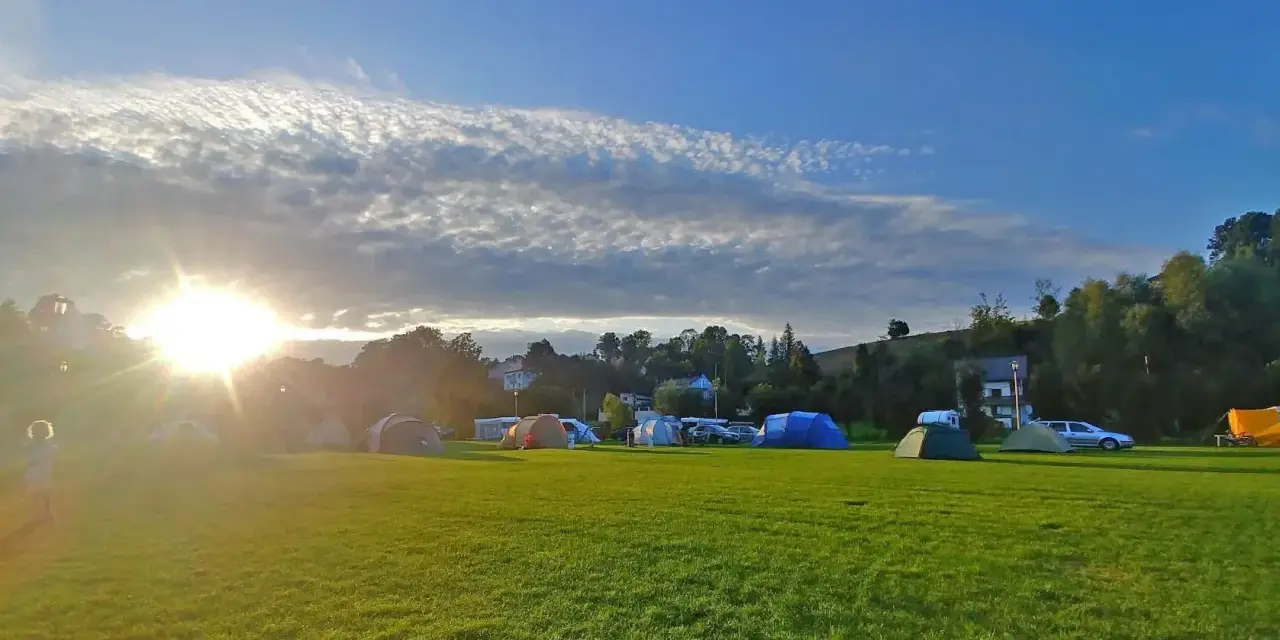 Słońce przebija się przez chmury nad polem namiotowym. Rozstawione namioty i samochody zapowiadają udany camping.