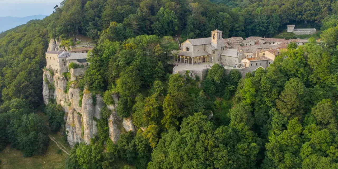 A monastery complex clings to a sheer cliff face in La Verna, Italy, surrounded by lush green forests.