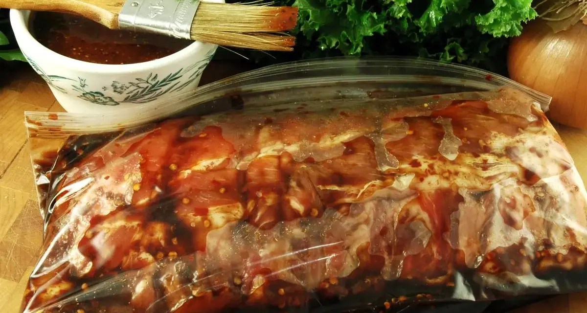 Karkówka w torebce, gotowa na grilla. W miseczce czeka aromatyczna marynata do karkówki na grilla, a obok pęczek zielonej sałaty i cebula.