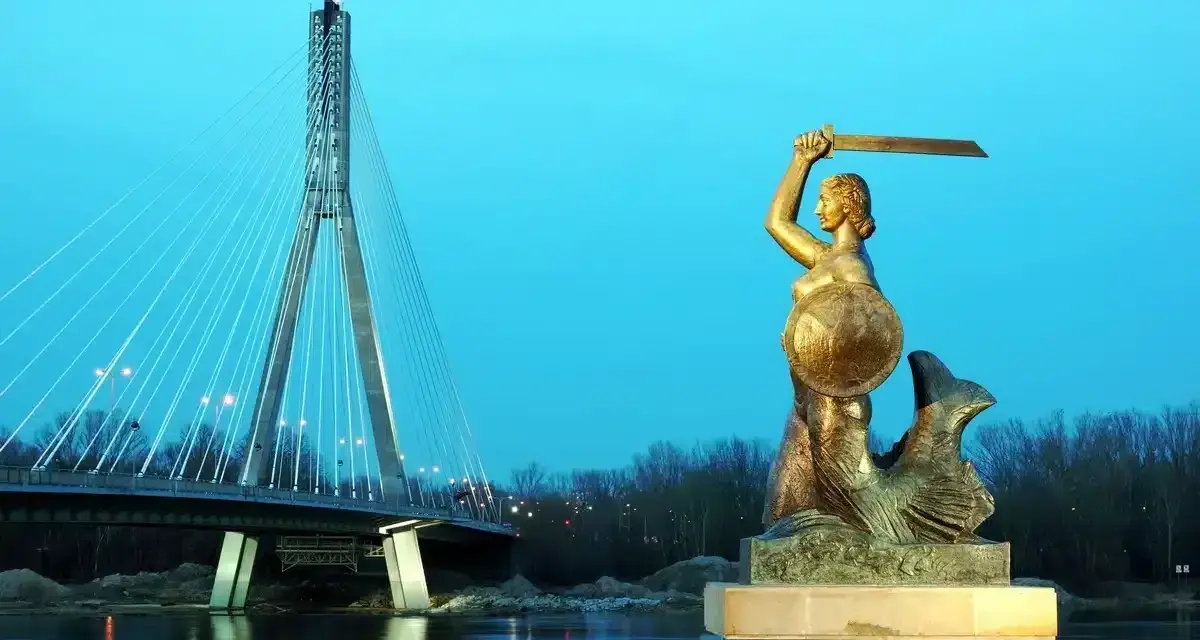 Syrenka warszawska z mieczem i tarczą stoi nad Wisłą, w tle nowoczesny most.
