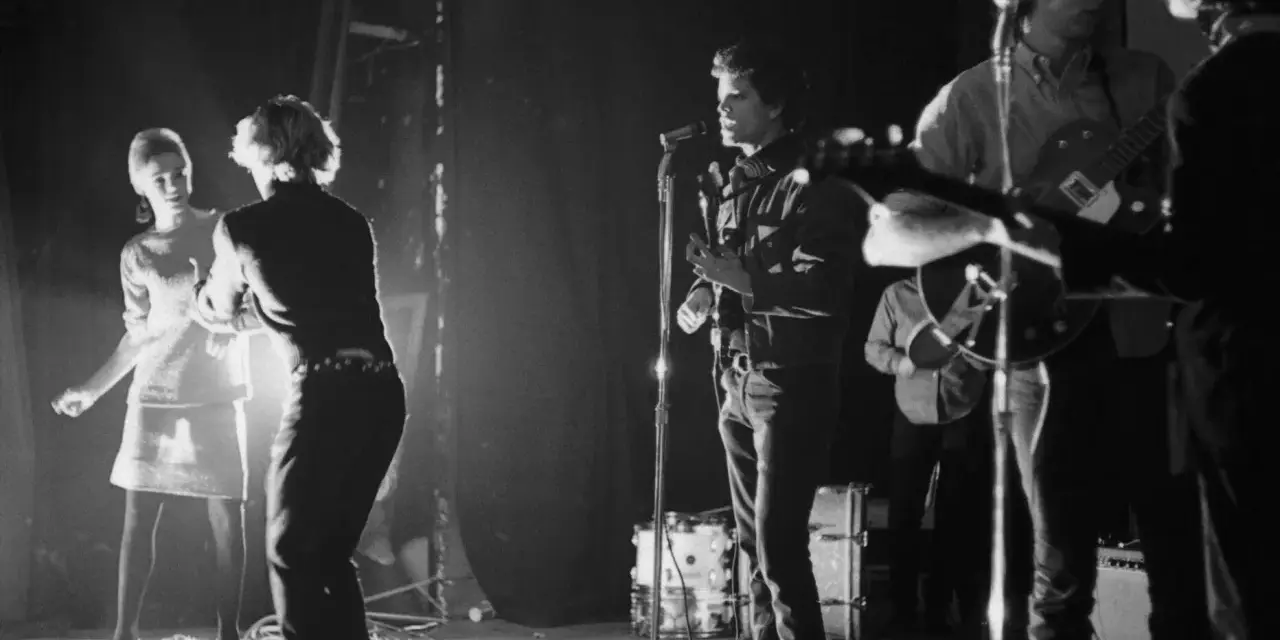 Scène de concert des membres de The Velvet Underground. Une femme danse à gauche, tandis que Lou Reed chante au micro.