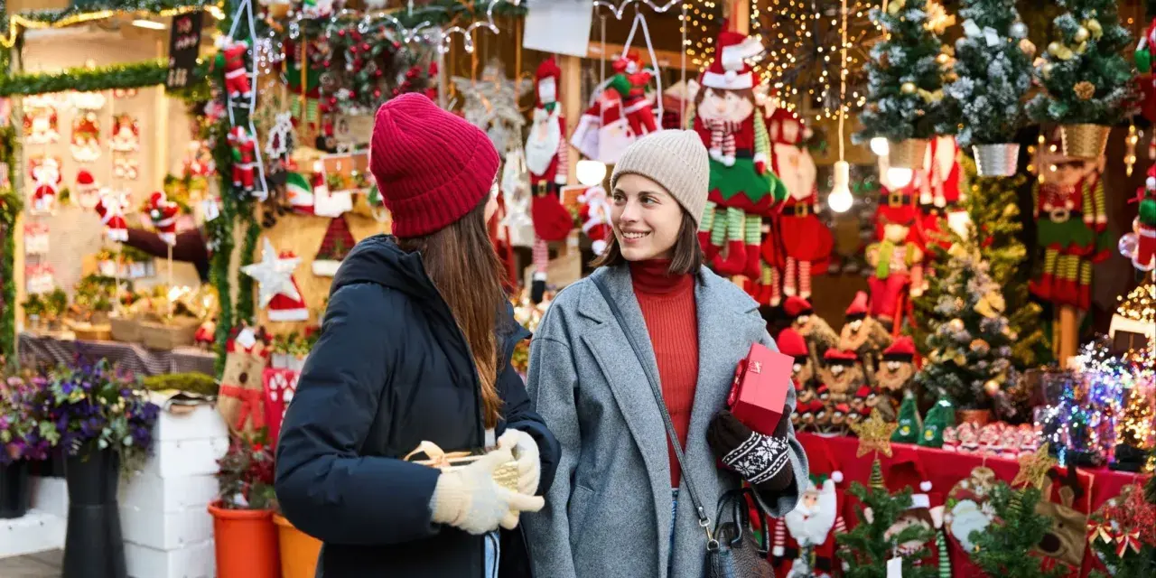 Två kvinnor njuter av julmarknader i Skåne, omgivna av julpynt och ljus. De håller presenter och ler mot varandra.