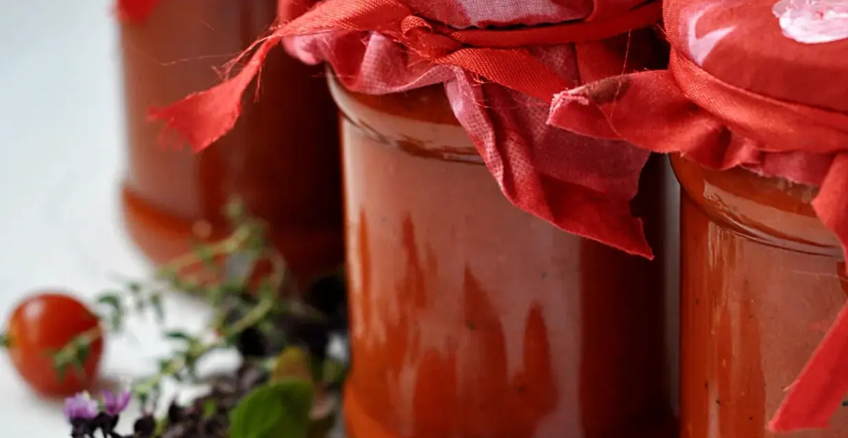 Słoiki z domowym sosem pomidorowym do makaronu, ozdobione czerwonymi chusteczkami. Obok świeże pomidorki i zioła.