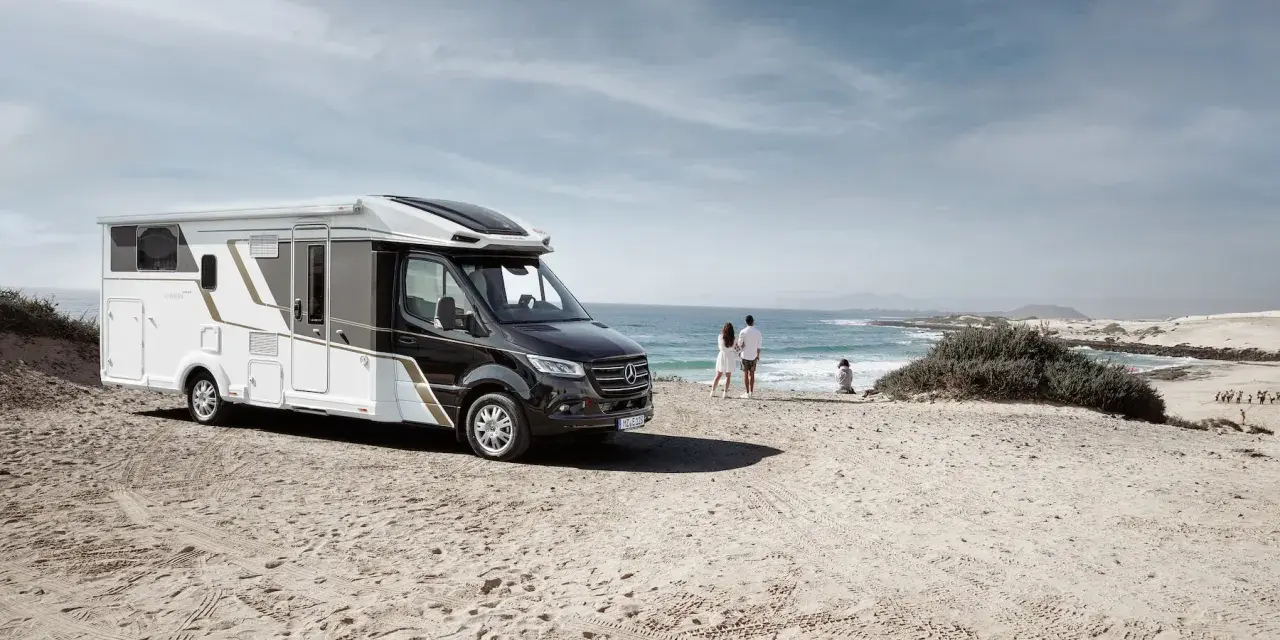 Eura Mobil husbil parkerad på en sandstrand vid havet. Ett par står och tittar ut över vågorna.