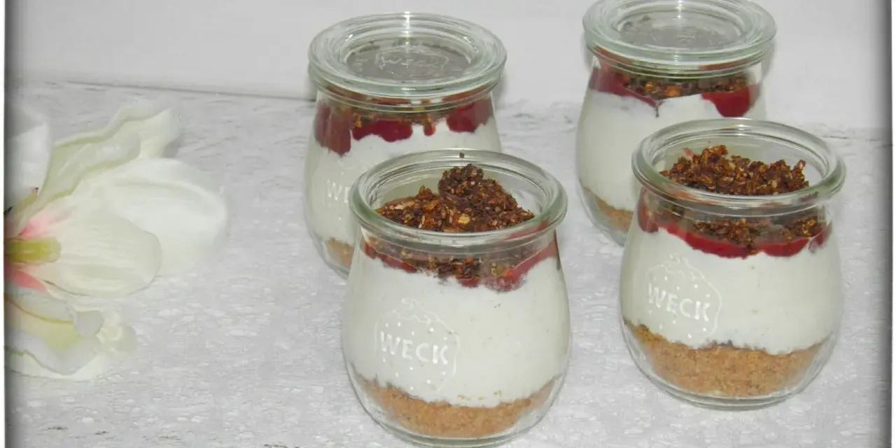 Vier leckere Desserts im Glas mit Krümelboden, cremiger Füllung, roter Fruchtsauce und knusprigem Topping.