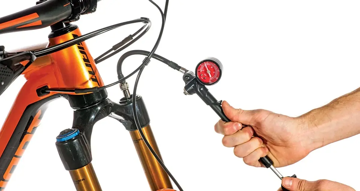 Adjusting the mtb suspension setup. A hand uses a shock pump with a gauge to set the air pressure on an orange mountain bike's front fork.