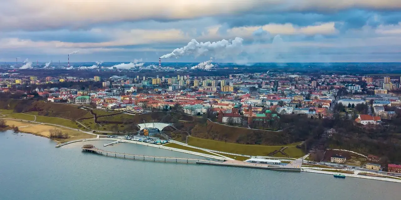Widok na Płock z lotu ptaka, z Wisłą w tle. Widać panoramę miasta, fabryki z dymiącymi kominami i nowoczesną infrastrukturę nad rzeką, która może być częścią atrakcji dla dzieci w Płocku.