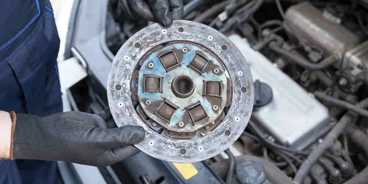 Mechanik w czarnych rękawiczkach trzyma zużyte sprzęgło samochodowe na tle silnika.