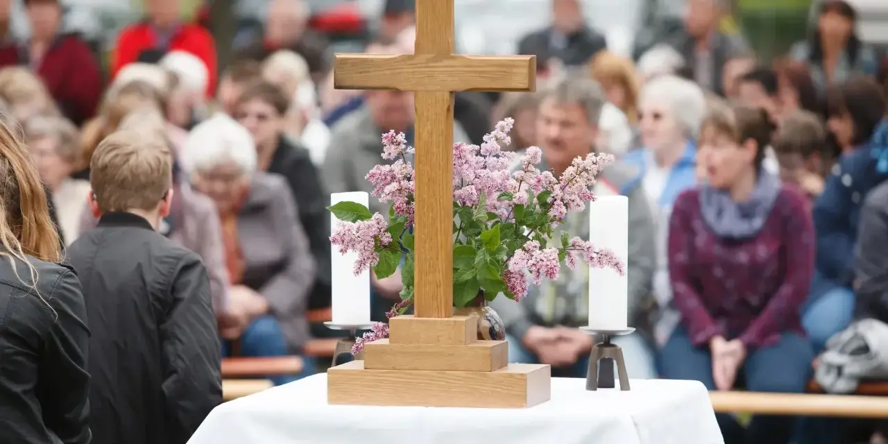 Holzkreuz mit Blumen und Kerzen vor einer Gemeinde. Ein Thema, das auch bei unterschiedlichen Konfessionen von Ehepartnern relevant ist: die Kirchensteuer.