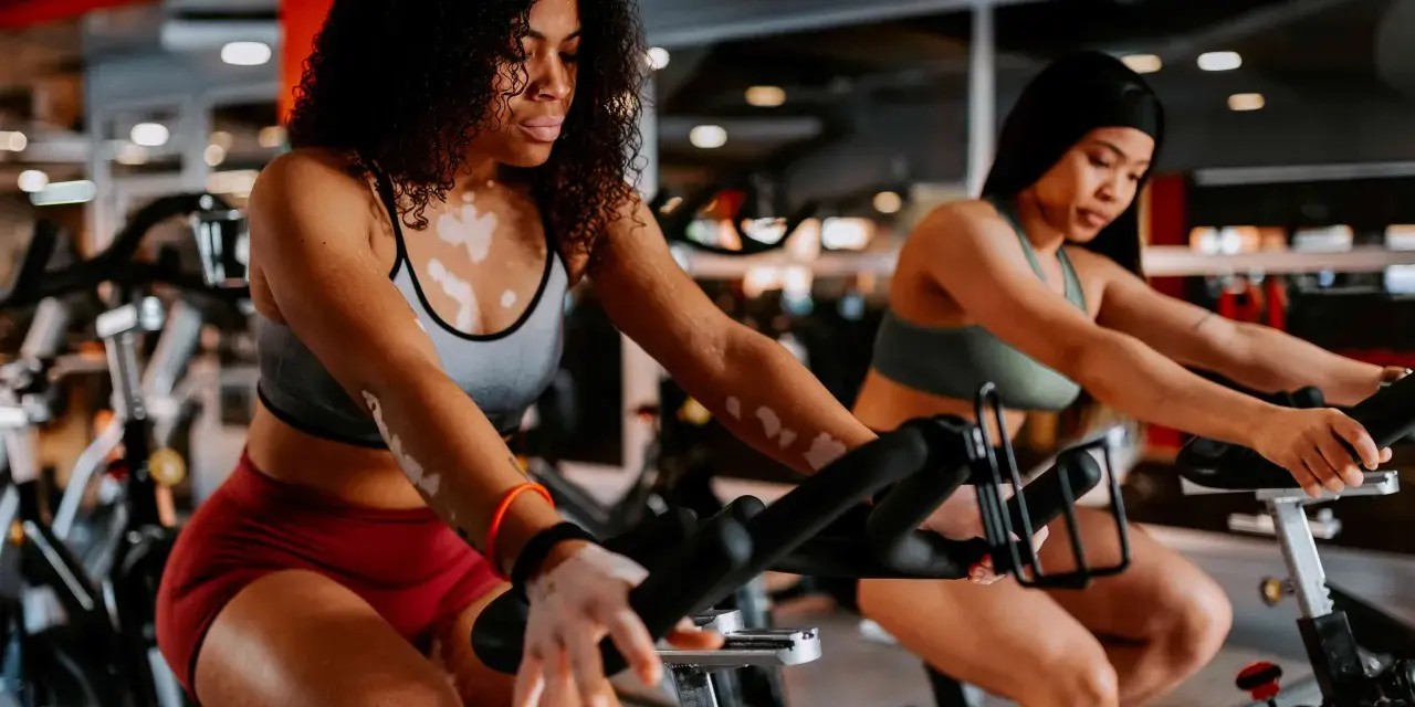 Two women are engaged in a stationary bike workout in a gym. The woman in the foreground has vitiligo and curly hair.