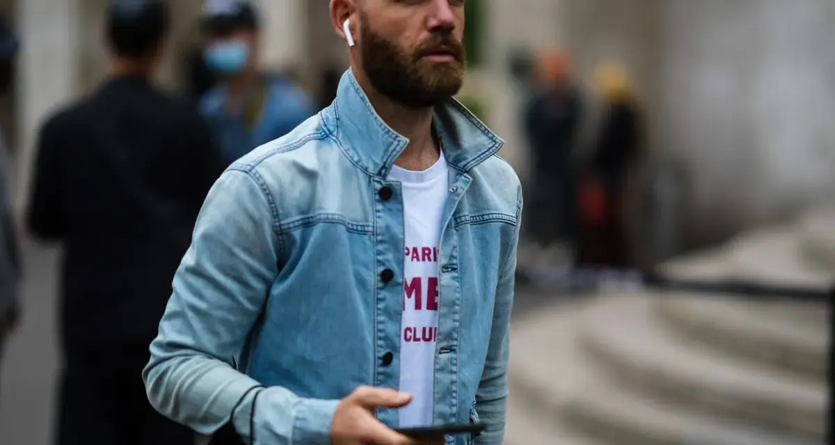 Hombre calvo sin barba, con auriculares y chaqueta vaquera, mira su móvil.