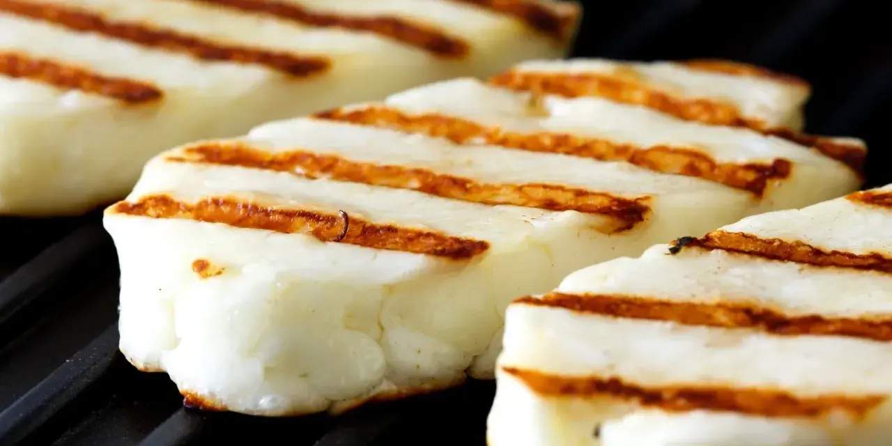 Trzy kawałki halloumi z grilla, z charakterystycznymi brązowymi paskami, leżą na czarnej, grillowej powierzchni.