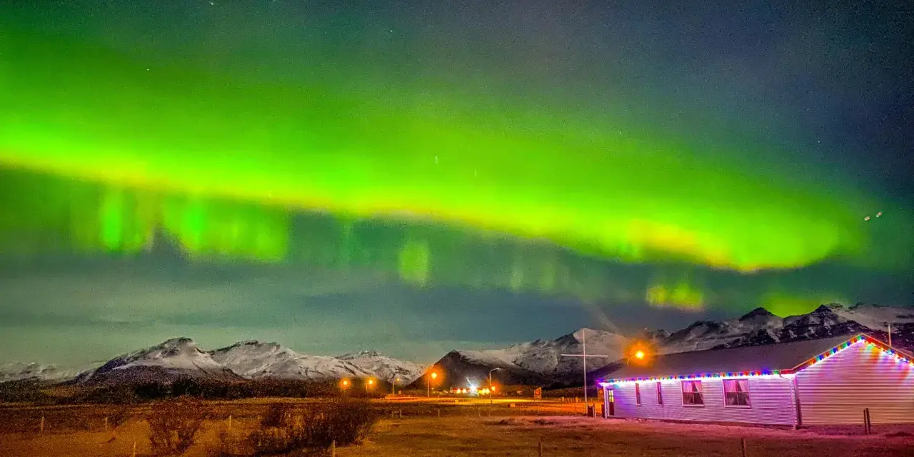 Islandia pogoda listopad: zorza polarna nad ośnieżonymi górami i domkiem z lampkami.