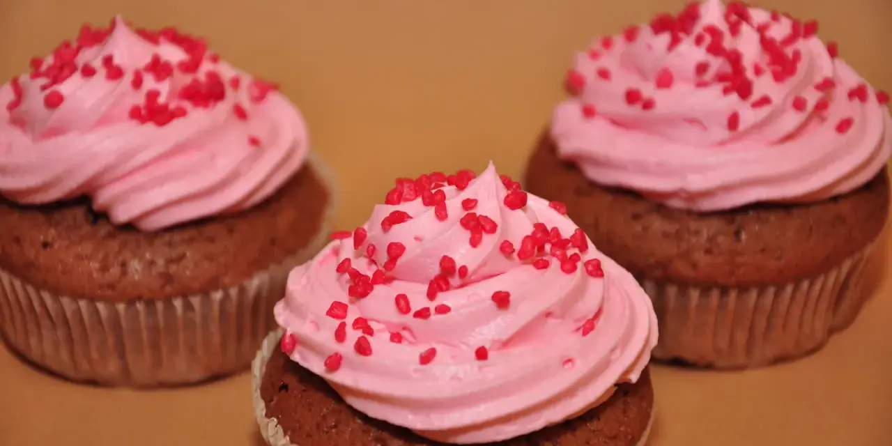 Tre chokladmuffins med rosa frosting och röda strössel, bakade i rosa muffinsformar.