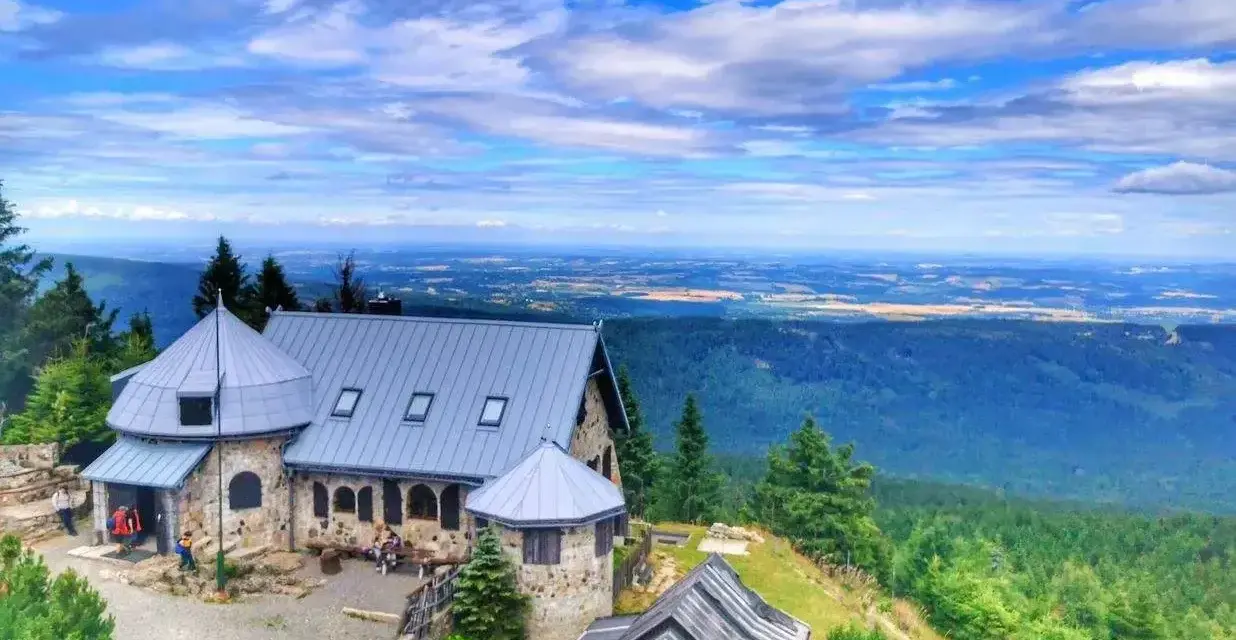 Kamienny schronisko z szarym dachem w Górach Izerskich, otoczone zielonymi lasami i rozległą panoramą dolin pod błękitnym, chmurowym niebem.