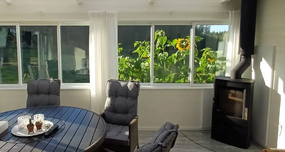 Mysig kamin i uterummet med solrosor utanför fönstret. Ett runt bord med stolar står redo för fika.